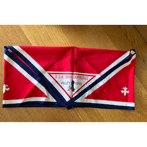 May include: A red, white, and blue BSA 1964 Jamboree neckerchief. The neckerchief has a white triangle with the text "B.S.A. 1964 JAMBOREE VALLEY FORGE" and a fleur-de-lis symbol. The neckerchief is folded.