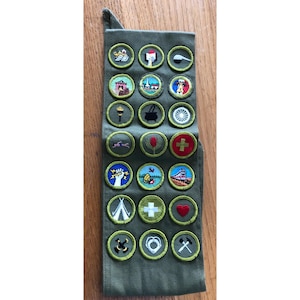 May include: A collection of embroidered patches, each with a unique design, arranged on a green fabric strip. The patches feature various symbols, including a torch, a heart, and a cross. The patches are round with a yellow border.