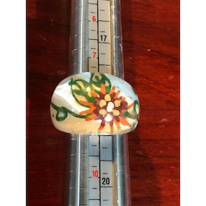 May include: A clear glass ring with a painted floral design. The flower is orange, brown, and white with green leaves.