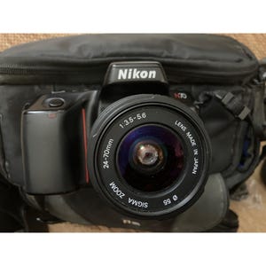 Op de afbeelding: Zwarte Nikon N70 camera met een Sigma Zoom lens. De lens heeft de specificaties 24-70mm, 1:3.5-5.6 en een diameter van 55mm. De camera staat tegen een zwarte cameratas.