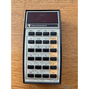 Puede incluir: Una taza de cerámica blanca con asa negra y el texto "But First, Coffee" en negrita negra. La taza se encuentra sobre un fondo borroso, resaltando su diseño limpio.