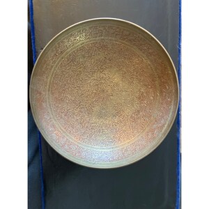 Pode incluir: Uma tigela redonda de latão antigo com intrincados padrões florais e de folhas gravados em sua superfície. A tigela tem uma borda ligeiramente elevada e um tom dourado quente, sugerindo idade e artesanato. O diâmetro é de cerca de 25 cm.