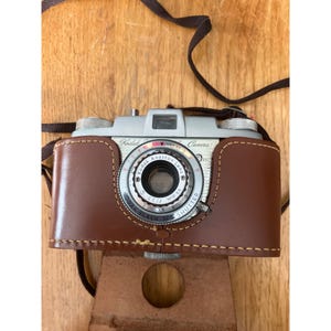 May include: Vintage Kodak camera with a brown leather case. The camera has a silver body with black and red details. The lens is labeled "Kodak Anastigmat Lens 44mm f/3.5". The case has a brown strap.