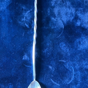Può includere: Un agitatore per cocktail in metallo color argento caratterizzato da un lungo manico sottile con un distinto design a spirale ritorta. La parte inferiore dell'agitatore termina con una piccola coppa ovale appuntita con un intaglio centrale.