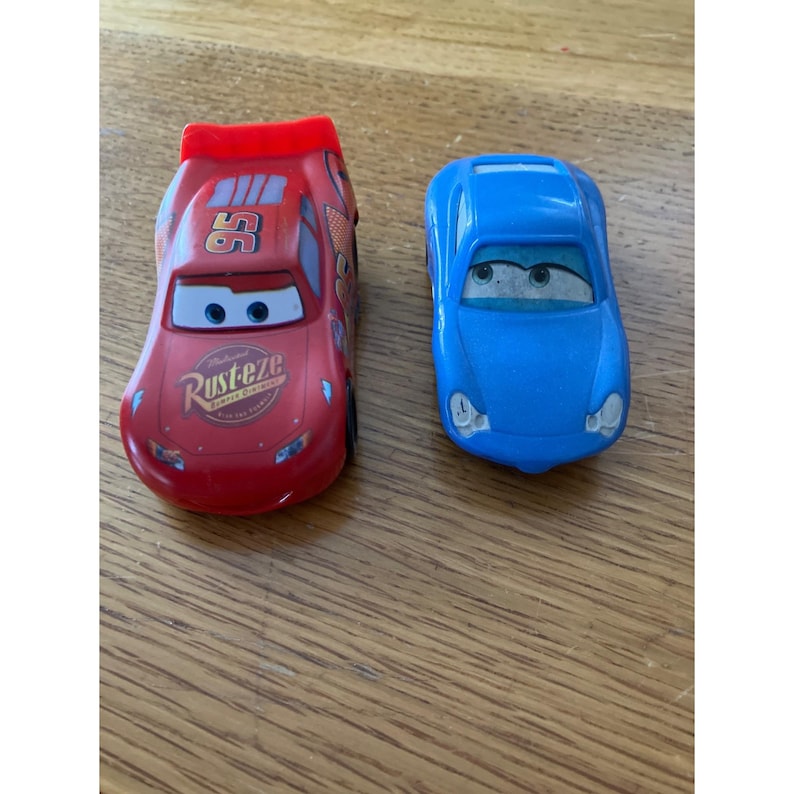 May include: Two toy cars featuring character designs. The red car displays the number 95, a Rust-eze logo, and a rear spoiler. The blue car has a smooth, rounded body with large windshield eyes. Both vehicles have painted eyes on the windshields.