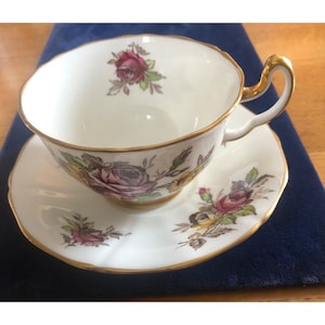 Peut inclure: Service à thé blanc avec une rose fleurie rose, jaune et verte. La tasse et la soucoupe ont une bordure dorée. La tasse a une poignée incurvée.