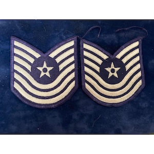 Könnte beinhalten: Zwei blau-weiße Militärabzeichen. Jedes Abzeichen hat einen blauen Hintergrund mit weißen Streifen und einem zentralen Sternemblem. Wahrscheinlich handelt es sich um militärische Dienstgradabzeichen.