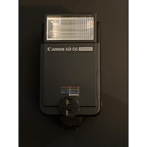 May include: A black Canon AB-56 Automatic flash unit. The flash has a rectangular head with a ribbed front. The Canon logo and model number are visible. The unit has a manual mode and distance settings.
