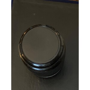 Puede incluir: Un objetivo de c&aacute;mara negro visto desde el frente, con una tapa circular que cubre el elemento de cristal. El cuerpo cil&iacute;ndrico del objetivo presenta una secci&oacute;n de agarre texturizada y una carcasa exterior lisa.