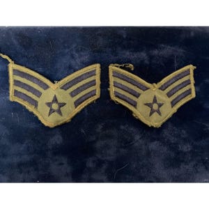 Pode incluir: Dois galões de patente militar em verde-oliva com três listras horizontais azul-escuras e uma estrela central azul-escura. Estes emblemas bordados têm formato em V com bordas costuradas, exibindo um design de insígnia clássico.