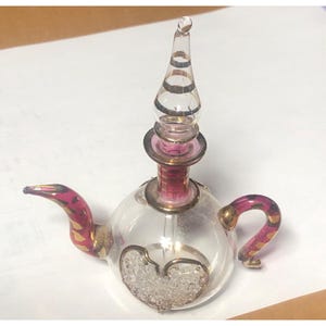 Può includere: Un flacone di profumo a forma di teiera in vetro trasparente con un design a forma di cuore. La bottiglia presenta un manico e un beccuccio rosa e oro, e un tappo decorativo con accenti dorati. La bottiglia è probabilmente vintage o antica.
