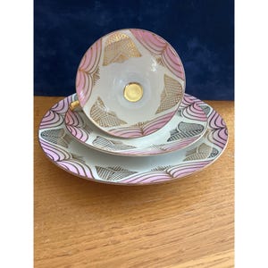 May include: A white porcelain teacup and saucer set featuring a repeating pattern of pink curved lines and gold geometric accents. The teacup has a solid gold center at the base and a gold-trimmed handle, while the saucer displays matching pink and gold motifs.