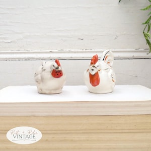May include: Two ceramic chicken figurines. One is a hen, the other a rooster. Both are white with red combs and beaks. The hen is squat, while the rooster stands upright. They are on a stack of books.