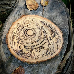 Peut inclure: Une tranche de bois ronde avec un motif de pyrogravure. L'œuvre présente une spirale centrale avec un croissant de lune, entourée d'éléments botaniques et de champignons. La pièce est de couleur bois naturel avec des détails marron foncé.