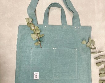 Puede incluir: Bolso tote de lino verde azulado claro con asas largas y un bolsillo delantero. El bolso tiene pequeños remaches plateados en el borde superior. Una ramita de eucalipto está metida en el bolsillo. Una pequeña etiqueta blanca con un gráfico es visible.