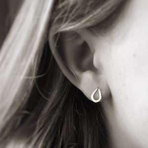 May include: A close-up of a person's ear with a small, silver teardrop-shaped earring. The earring is simple and delicate.