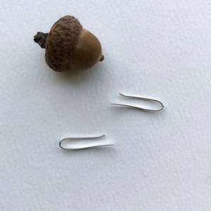 Puede incluir: Dos ganchos de pendientes plateados sobre una superficie blanca con una bellota marr&oacute;n.