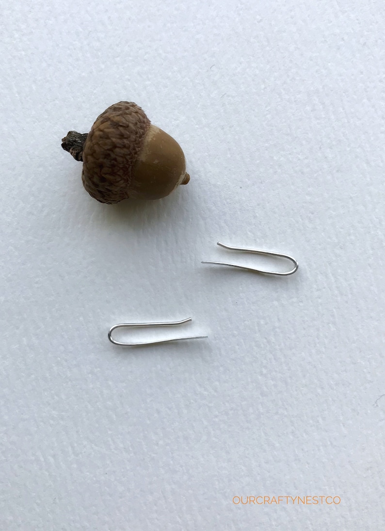 Puede incluir: Dos ganchos de pendientes plateados sobre una superficie blanca con una bellota marr&oacute;n.