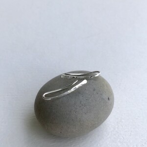 Puede incluir: Un par de pendientes de aro martillados de plata sobre una piedra gris. Los pendientes son simples y delicados, con una forma ligeramente curvada.