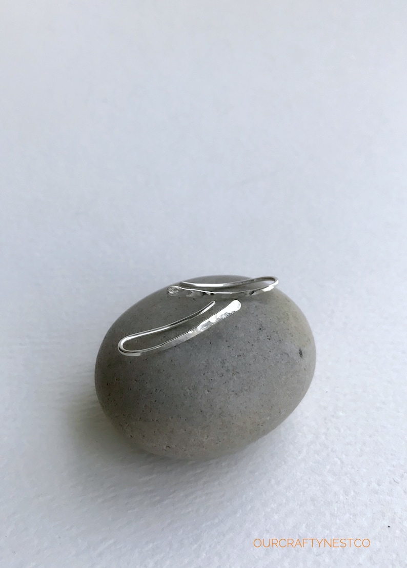Puede incluir: Un par de pendientes de aro martillados de plata sobre una piedra gris. Los pendientes son simples y delicados, con una forma ligeramente curvada.