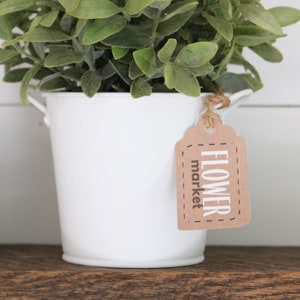 Puede incluir: Un cubo de metal blanco con una planta verde de hojas dentro. El cubo tiene una etiqueta marrón atada al asa que dice "FLOWER market".