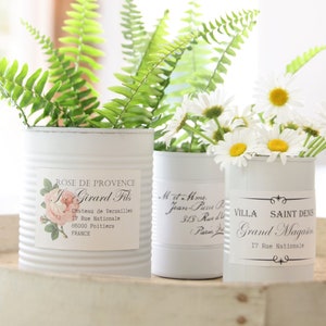 May include: Three white tin cans repurposed as vases, each holding greenery and flowers. The cans have labels with French text and floral designs. The vases sit on a light-colored, oval tray.