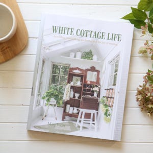 May include: A book titled "White Cottage Life" with the subtitle "decorating, repurposing, gardening, and the joy of creating." The cover features a detailed interior scene with a wooden cabinet, plants, and white accents. The book is on a white wooden surface.