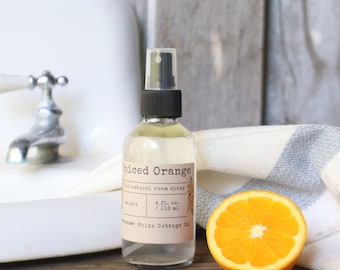 May include: A clear glass bottle of "Spiced Orange" all-natural room spray with a black spray nozzle. The label includes the text "4 fl. oz. / 118 ml." A sliced orange and a striped towel are next to the bottle.