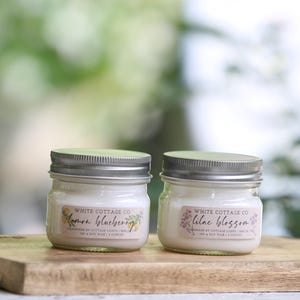 Puede incluir: Dos pequeños tarros de velas de vidrio con tapas de metal sobre una tabla de madera. Las etiquetas dicen "Lemon Blueberry" y "Lilac Blossom" con ilustraciones florales. El fondo es verde suave.