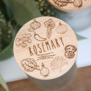 K&ouml;nnte beinhalten: Nahaufnahme eines Holzdeckels mit dem Wort "ROSEMARY" in der Mitte eingraviert. Der Deckel ist mit Illustrationen verschiedener Kr&auml;uter und Gew&uuml;rze verziert, darunter Knoblauch, Ingwer und Zwiebel. Der Deckel befindet sich auf einem Glas.