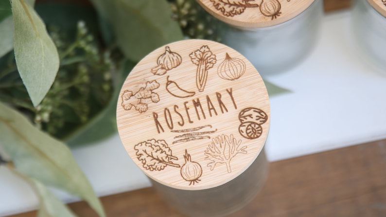K&ouml;nnte beinhalten: Nahaufnahme eines Holzdeckels mit dem Wort "ROSEMARY" in der Mitte eingraviert. Der Deckel ist mit Illustrationen verschiedener Kr&auml;uter und Gew&uuml;rze verziert, darunter Knoblauch, Ingwer und Zwiebel. Der Deckel befindet sich auf einem Glas.