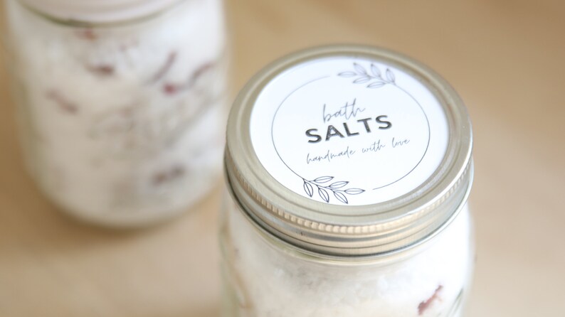 May include: Two glass jars filled with white bath salts, with silver lids. The jar in the foreground has a white label that reads "bath salts handmade with love".