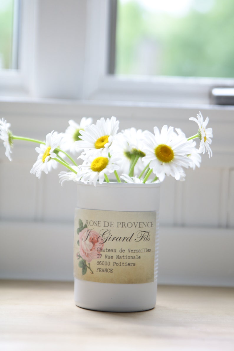 May include: A white ceramic vase with a label that reads "ROSE DE PROVENCE I. Girard Fils Chateau de Versailles 17 Rue Nationale 86000 Poitiers FRANCE" filled with white daisies.