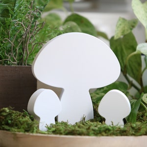 Puede incluir: Una composición decorativa con tres recortes de hongos blancos de varios tamaños, colocados en medio de musgo verde y vegetación. El fondo incluye plantas en macetas con exuberante follaje verde, creando una escena natural y caprichosa.