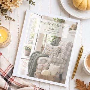 May include: A softcover book titled White Cottage Fall by Mary Yoder, featuring a cover image of a plaid armchair with a green throw blanket, a floral pillow, and a basket of pumpkins, displayed on a white wooden surface with a gold pen.
