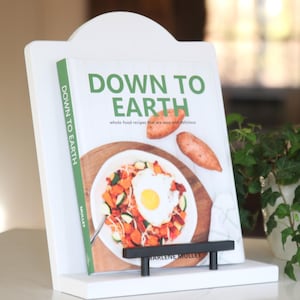 以下が含まれることがあります： マルリーン・マレット著「ダウン・トゥ・アース」というタイトルの料理本が置かれた白い料理本スタンド。料理本には、サツマイモ、ズッキーニ、卵が入ったボウルに入った料理の写真が掲載されています。