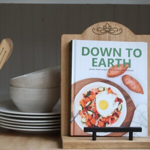 K&ouml;nnte beinhalten: Ein h&ouml;lzerner Kochbuchst&auml;nder h&auml;lt ein Kochbuch mit dem Titel "DOWN TO EARTH". Das Cover zeigt ein farbenfrohes Gericht mit Spiegelei und S&uuml;&szlig;kartoffeln. Eine wei&szlig;e Kanne mit Holzl&ouml;ffeln und ein Stapel Teller und Sch&uuml;sseln befinden sich links.