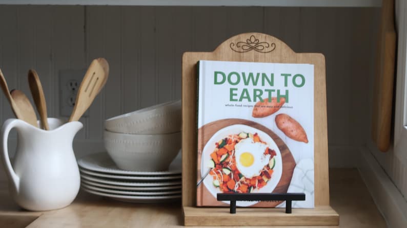 K&ouml;nnte beinhalten: Ein h&ouml;lzerner Kochbuchst&auml;nder h&auml;lt ein Kochbuch mit dem Titel "DOWN TO EARTH". Das Cover zeigt ein farbenfrohes Gericht mit Spiegelei und S&uuml;&szlig;kartoffeln. Eine wei&szlig;e Kanne mit Holzl&ouml;ffeln und ein Stapel Teller und Sch&uuml;sseln befinden sich links.
