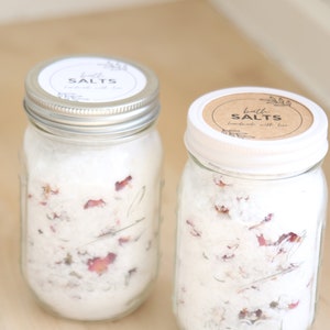 May include: Two glass jars filled with white bath salts and dried rose petals. The jars have white lids with brown paper labels that read "bath salts" in black text.
