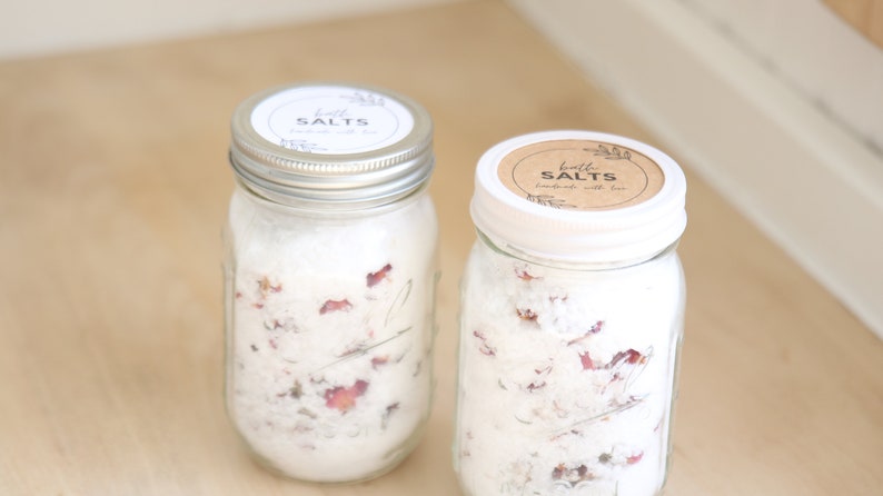 May include: Two glass jars filled with white bath salts and dried rose petals. The jars have white lids with brown paper labels that read "bath salts" in black text.