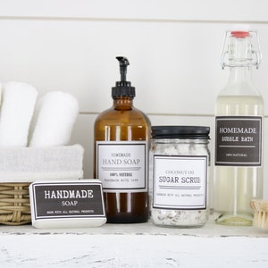 Op de afbeelding: Een verzameling badkamerproducten, waaronder een bruine glazen fles met een pompdispenser met het label "Homemade Hand Soap", een heldere glazen fles met een metalen deksel met het label "Homemade Bubble Bath", een glazen pot met een zwart deksel met het label "Coconut Oil Sugar Scrub", een stuk zeep met het label "Handmade Soap" en een houten borstel.