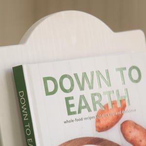 K&ouml;nnte beinhalten: Ein wei&szlig;er Kochbuchst&auml;nder aus Holz, der ein Kochbuch mit dem Titel "DOWN TO EARTH" in gr&uuml;nen Buchstaben h&auml;lt. Das Buch enth&auml;lt Vollwertkost-Rezepte und Bilder von S&uuml;&szlig;kartoffeln und anderen Zutaten.