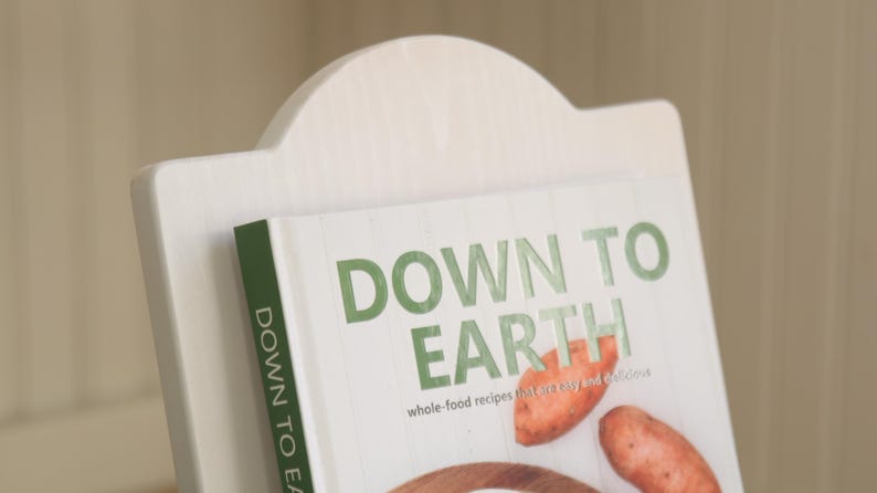 K&ouml;nnte beinhalten: Ein wei&szlig;er Kochbuchst&auml;nder aus Holz, der ein Kochbuch mit dem Titel "DOWN TO EARTH" in gr&uuml;nen Buchstaben h&auml;lt. Das Buch enth&auml;lt Vollwertkost-Rezepte und Bilder von S&uuml;&szlig;kartoffeln und anderen Zutaten.