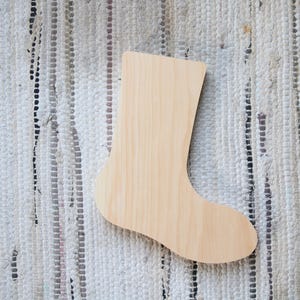 May include: A wooden stocking-shaped cutout, perfect for holiday crafts. The unfinished wood has a light, natural color. The cutout is placed on a textured, off-white rug with vertical stripes.