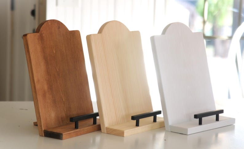 K&ouml;nnte beinhalten: Drei Tablet-St&auml;nder aus Holz in verschiedenen Farben: Dunkelbraun, Naturholz und Wei&szlig;. Jeder St&auml;nder hat eine rechteckige Basis mit einem schwarzen Balken zur Aufnahme eines Tablets und eine geschwungene Oberseite.