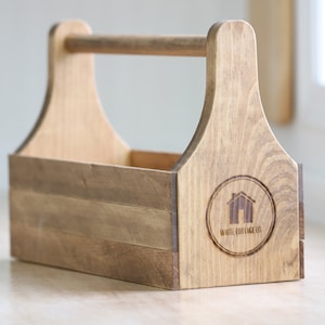 Op de afbeelding: Een lichtbruine houten gereedschapskist met een handvat. De kist heeft een rechthoekige basis en een handvat voor gemakkelijk dragen. Het White Cottage Co. logo is zichtbaar op de zijkant.