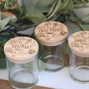K&ouml;nnte beinhalten: Drei transparente Gew&uuml;rzgl&auml;ser mit Holzdeckeln. Die Deckel sind mit den W&ouml;rtern "Rosmarin", "Chilipulver" und "Knoblauchsalz" sowie Illustrationen von Kr&auml;utern und Gem&uuml;se graviert. Die Gl&auml;ser werden auf einer Holzoberfl&auml;che mit gr&uuml;nem Laub im Hintergrund pr&auml;sentiert.