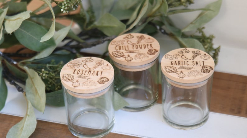 K&ouml;nnte beinhalten: Drei transparente Gew&uuml;rzgl&auml;ser mit Holzdeckeln. Die Deckel sind mit den W&ouml;rtern "Rosmarin", "Chilipulver" und "Knoblauchsalz" sowie Illustrationen von Kr&auml;utern und Gem&uuml;se graviert. Die Gl&auml;ser werden auf einer Holzoberfl&auml;che mit gr&uuml;nem Laub im Hintergrund pr&auml;sentiert.