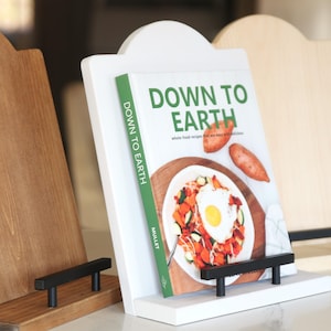 K&ouml;nnte beinhalten: Drei Kochbuchst&auml;nder aus Holz in verschiedenen Braun- und Wei&szlig;t&ouml;nen. Der mittlere St&auml;nder h&auml;lt ein Kochbuch mit dem Titel "Down to Earth" mit einem Foto eines Gerichts. Jeder St&auml;nder hat eine schwarze Metallst&uuml;tze.