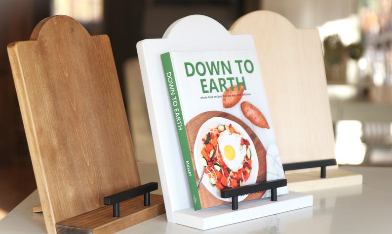 K&ouml;nnte beinhalten: Drei Kochbuchst&auml;nder aus Holz in verschiedenen Braun- und Wei&szlig;t&ouml;nen. Der mittlere St&auml;nder h&auml;lt ein Kochbuch mit dem Titel "Down to Earth" mit einem Foto eines Gerichts. Jeder St&auml;nder hat eine schwarze Metallst&uuml;tze.